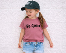 Load image into Gallery viewer, A young girl wearing a green baseball cap and a pink t-shirt with the text "Big Sister" stands against a white brick wall.
