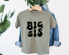 Load image into Gallery viewer, A gray t-shirt with the text "BIG SIS" printed on it, hanging on a wooden hanger.
