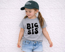 Load image into Gallery viewer, A young girl wearing a green baseball cap and a gray t-shirt with the text "BIG SIS" is leaning against a white wall.

