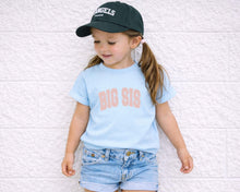 Load image into Gallery viewer, A young girl wearing a black baseball cap, a light blue t-shirt with the text "BIG SIS", and denim shorts is leaning against a white brick wall.
