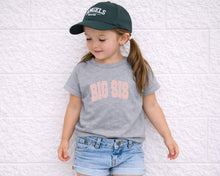 Load image into Gallery viewer, A young girl wearing a green baseball cap and a gray t-shirt with the text "BIG SIS" is leaning against a white brick wall.
